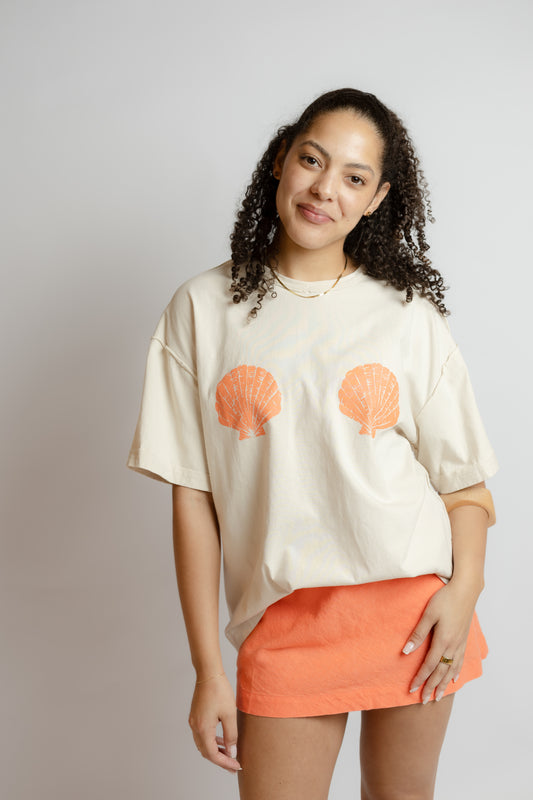 Woman wearing a cream t-shirt with shell designs and an orange skirt on a plain background