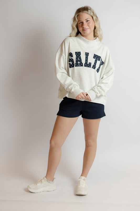 Person wearing a white sweatshirt with 'SALTY' text, black shorts, and white sneakers on a plain background