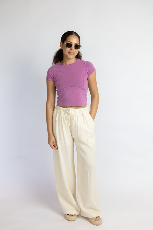 Woman wearing a purple top and white linen pants against a white background