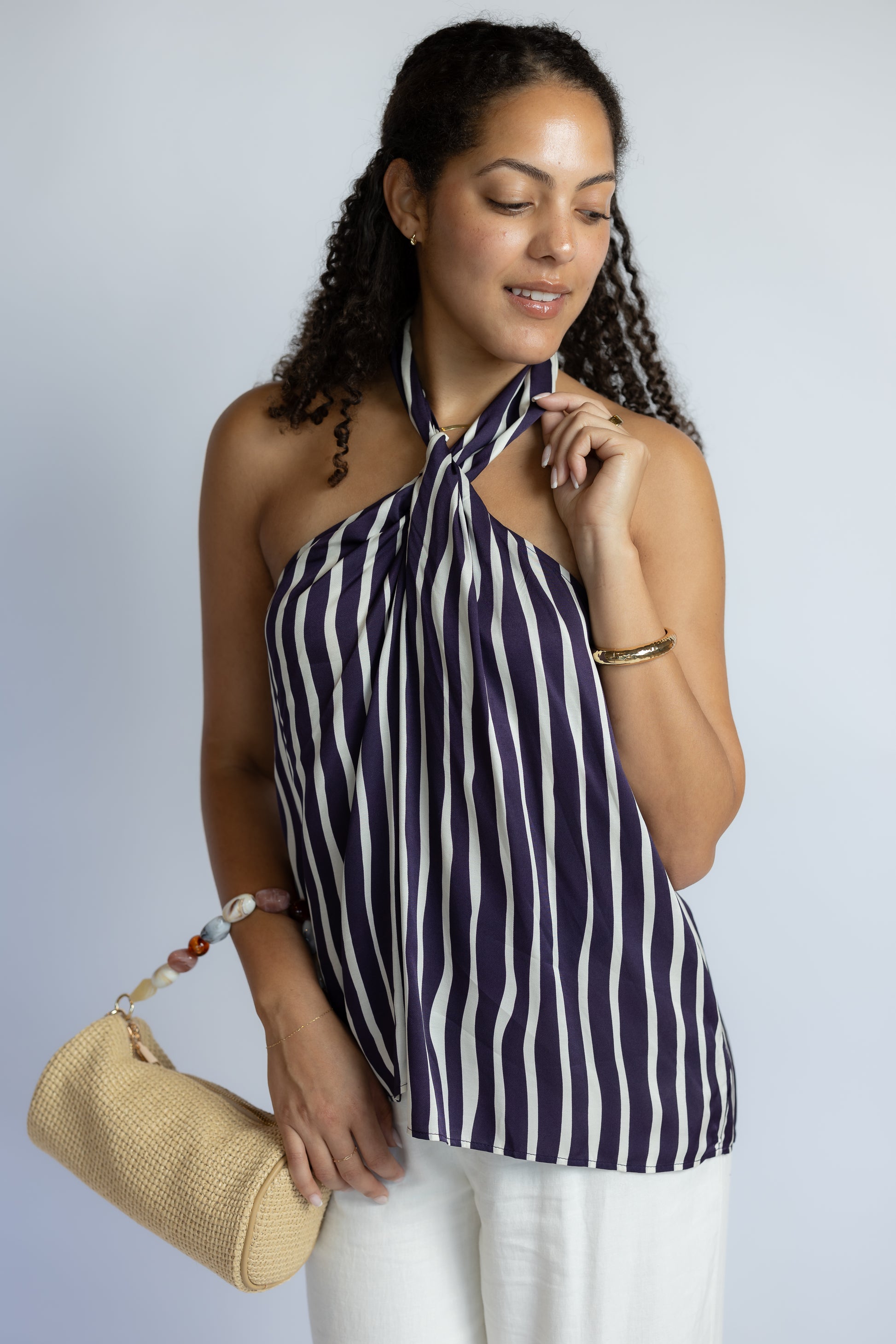 Model wearing the ASTR Cira striped halter top featuring a twisted neckline, open back, and flowy relaxed silhouette paired with white pants.
