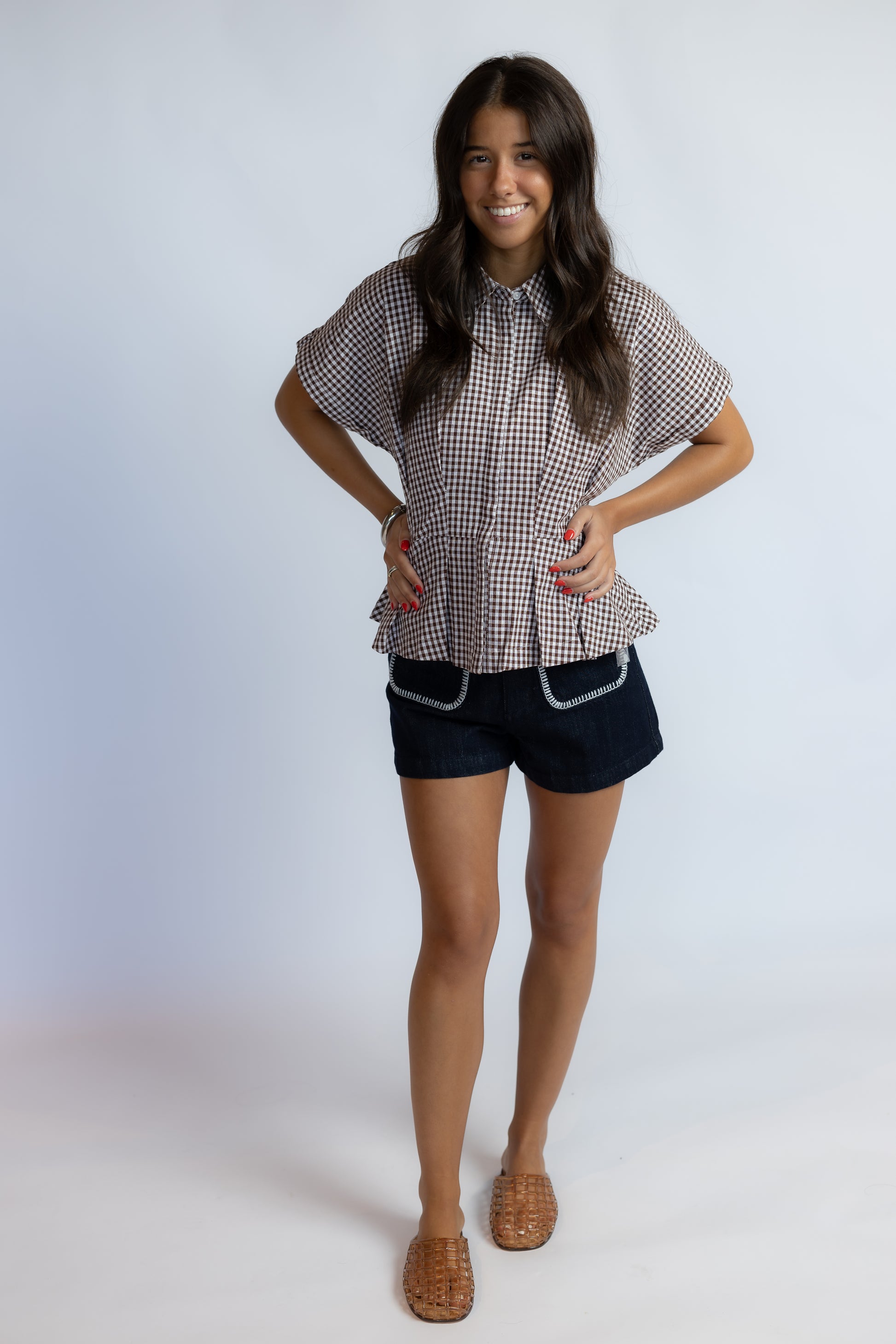 Model wearing the ASTR Beck Top, a brown and white gingham cotton blouse with pleated details, a concealed button-front closure, and rolled short sleeves.
