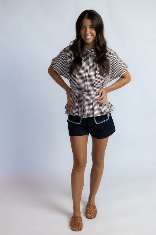 Model wearing the ASTR Beck Top, a brown and white gingham cotton blouse with pleated details, a concealed button-front closure, and rolled short sleeves.