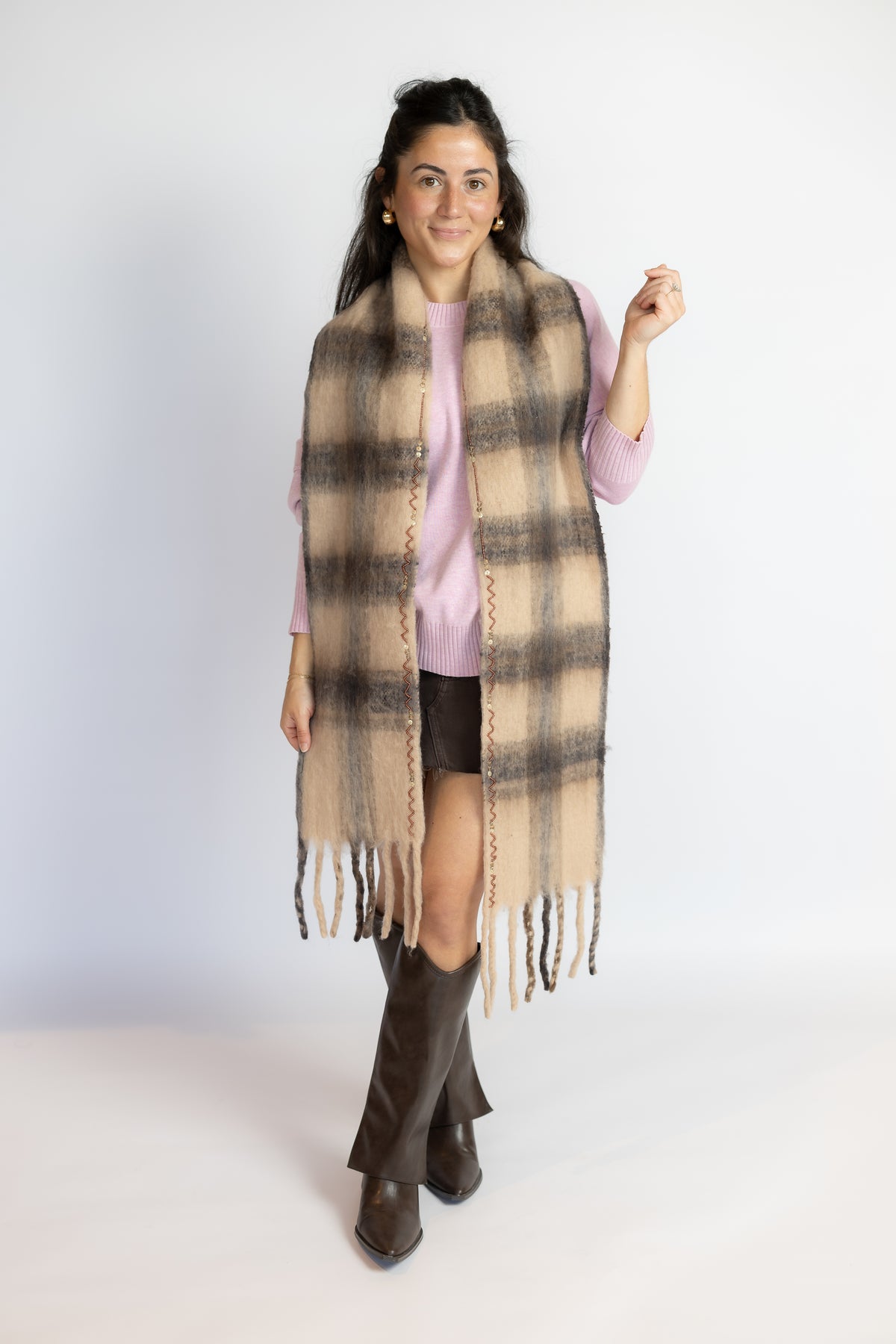 A woman modeling a long beige and brown plaid fringed scarf wrap over a light pink top and dark shorts, standing against a plain light gray studio background.