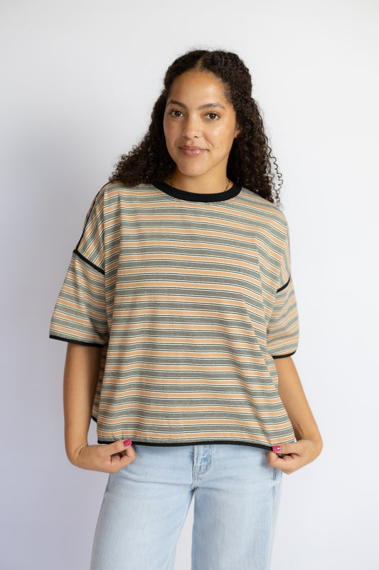A woman modeling a short-sleeve striped t-shirt with beige, brown, and black horizontal stripes, paired with light blue jeans, against a plain light gray background.