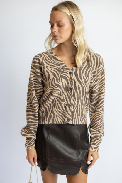A blonde woman wearing a beige and brown zebra-print long-sleeve button-up cardigan with a black faux-leather mini skirt, standing against a light gray studio background.