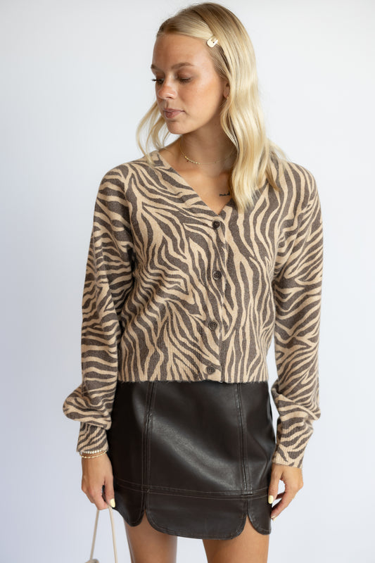 A blonde woman wearing a beige and brown zebra-print long-sleeve button-up cardigan with a black faux-leather mini skirt, standing against a light gray studio background.