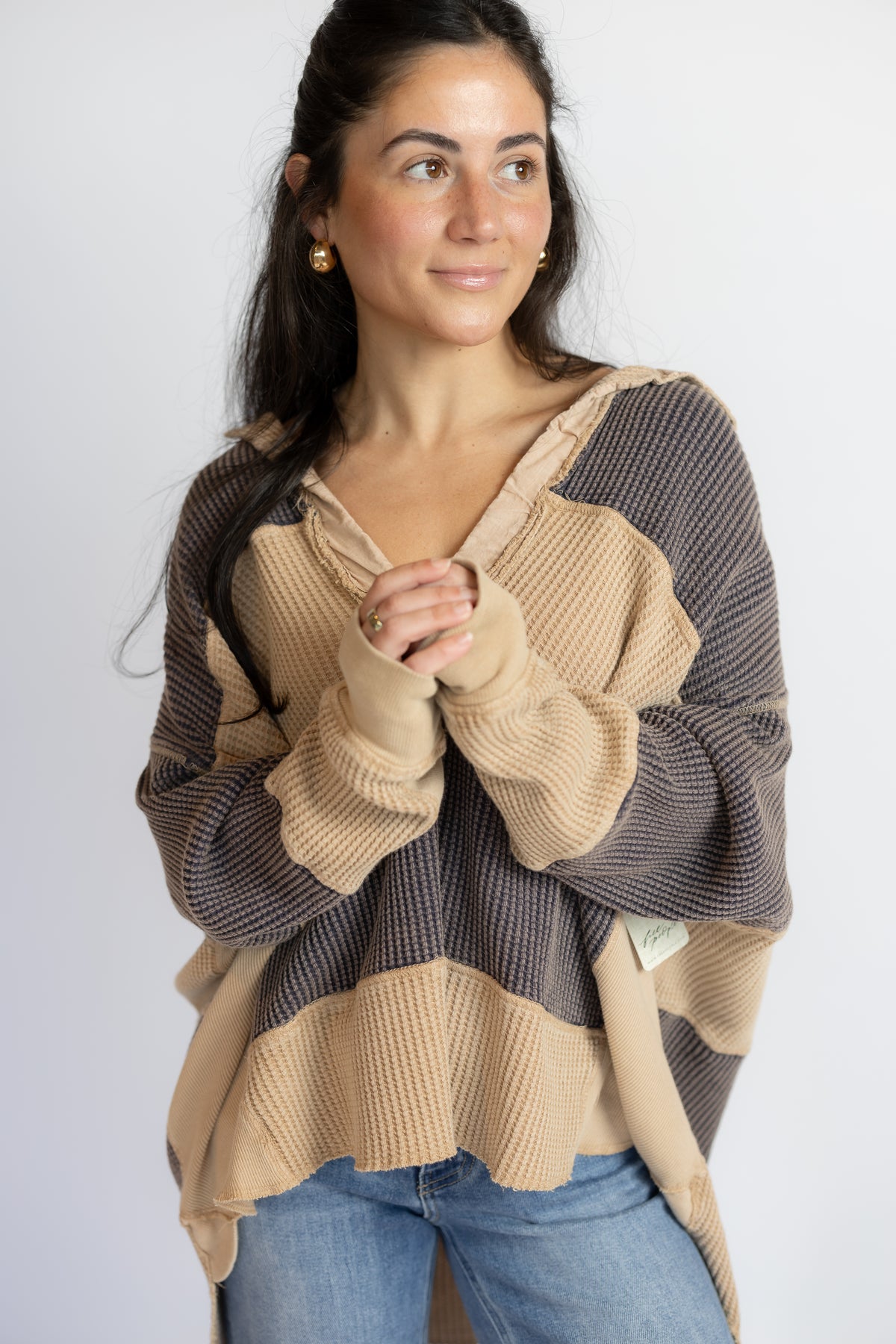 A woman wearing a waffle-knit color-block sweater in beige and dark brown, paired with blue jeans, against a plain light gray background.