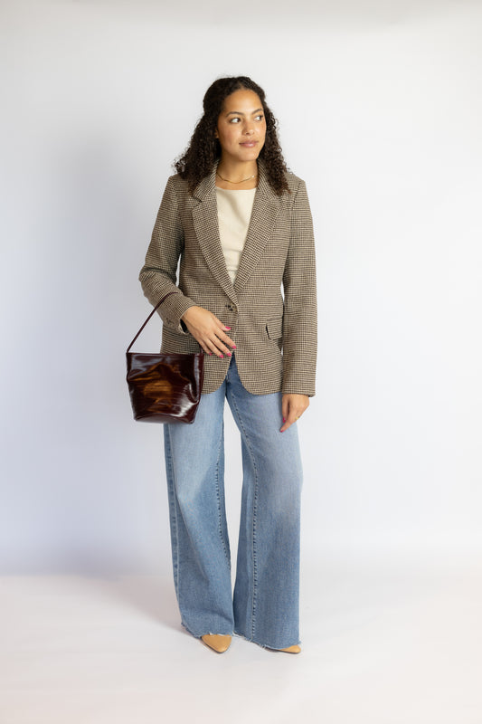 A woman stands against a light gray studio background wearing a taupe-brown textured blazer, light blue wide-leg jeans, a cream top, and holding a dark brown leather shoulder bag.