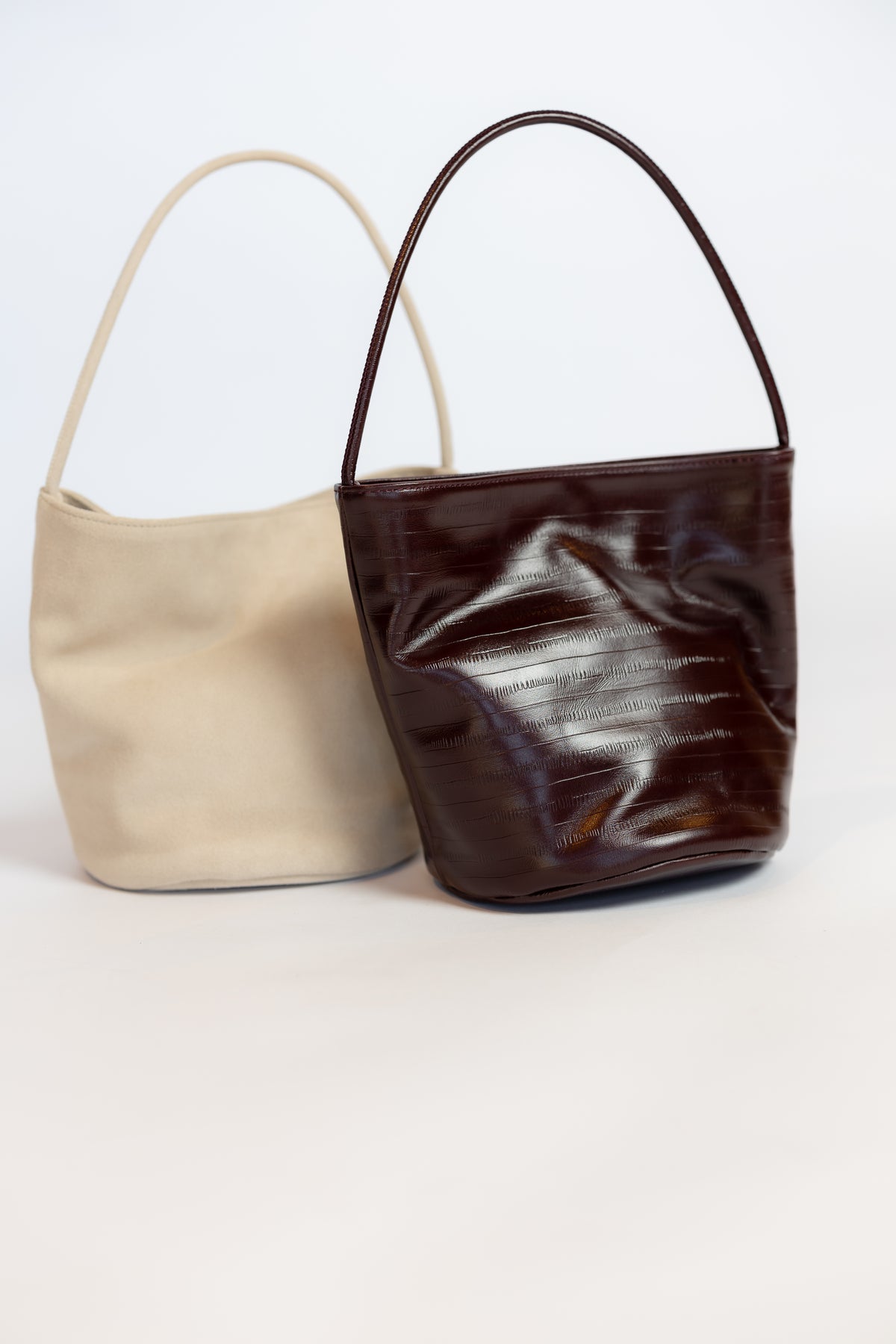 Two small shoulder handbags on a plain light background: one in smooth cream and one in glossy dark brown patent material, both with thin top handles.