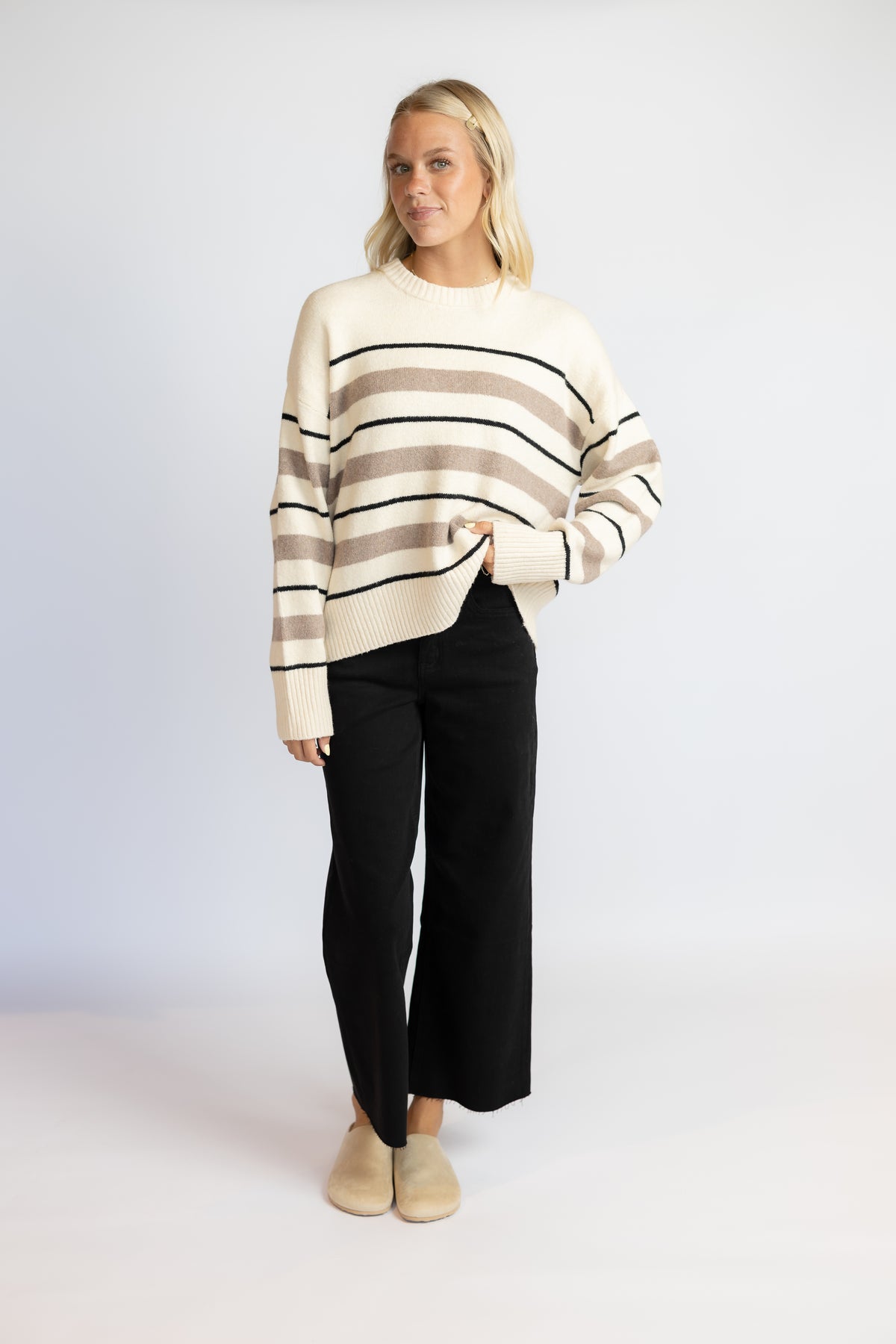 A woman modeling a cream knit sweater with beige and brown horizontal stripes, paired with black wide-leg pants and beige slip-on shoes, against a light gray studio background.