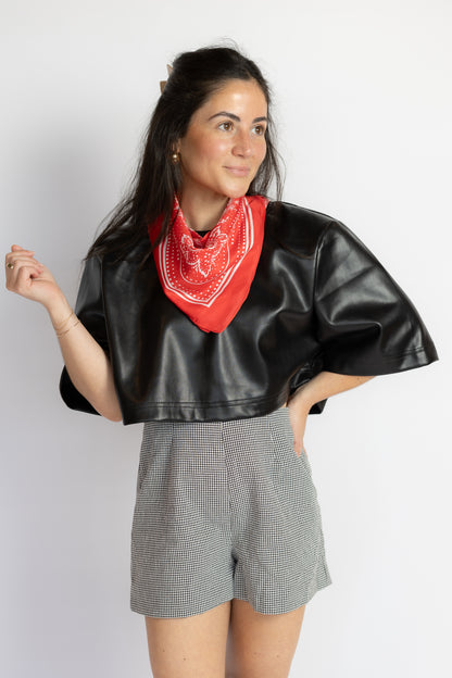 A woman modeling a black faux-leather cropped jacket with wide short sleeves, paired with gray houndstooth shorts and a red bandana, against a plain light gray background.