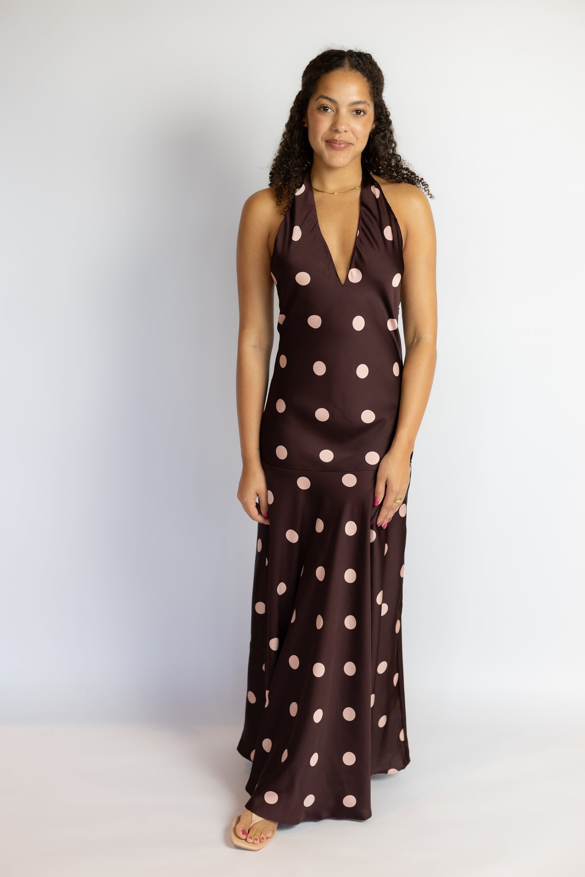 A woman modeling a dark brown halter-neck maxi dress with large light pink polka dots, standing against a plain light gray studio background.