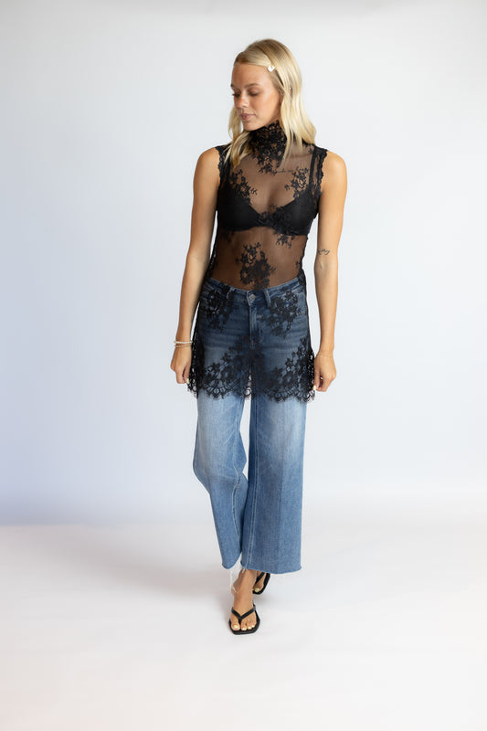 A woman modeling a sheer black lace mock-neck top paired with wide-leg blue jeans featuring black lace trim at the hem, against a plain light gray studio background.
