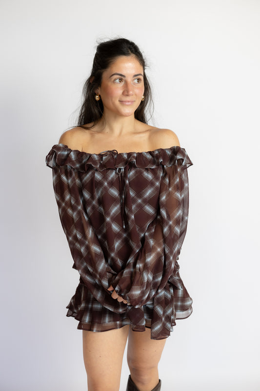 A woman modeling an off-the-shoulder long-sleeve blouse with a brown, black, and white plaid pattern and ruffled trim, set against a plain light gray background.