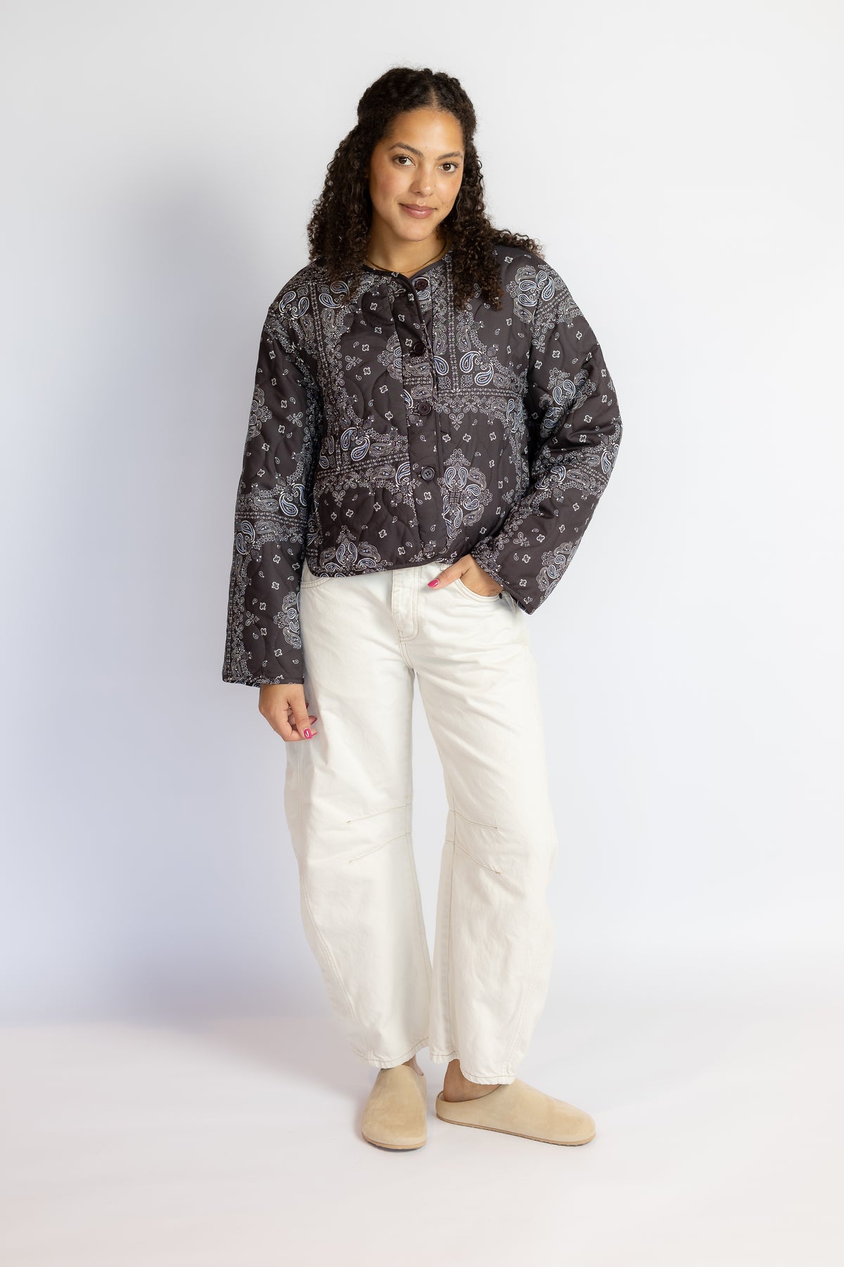 A woman modeling a dark gray long-sleeve cropped hoodie with a white paisley pattern, paired with white pants and beige slip-on shoes, standing against a light gray studio background.