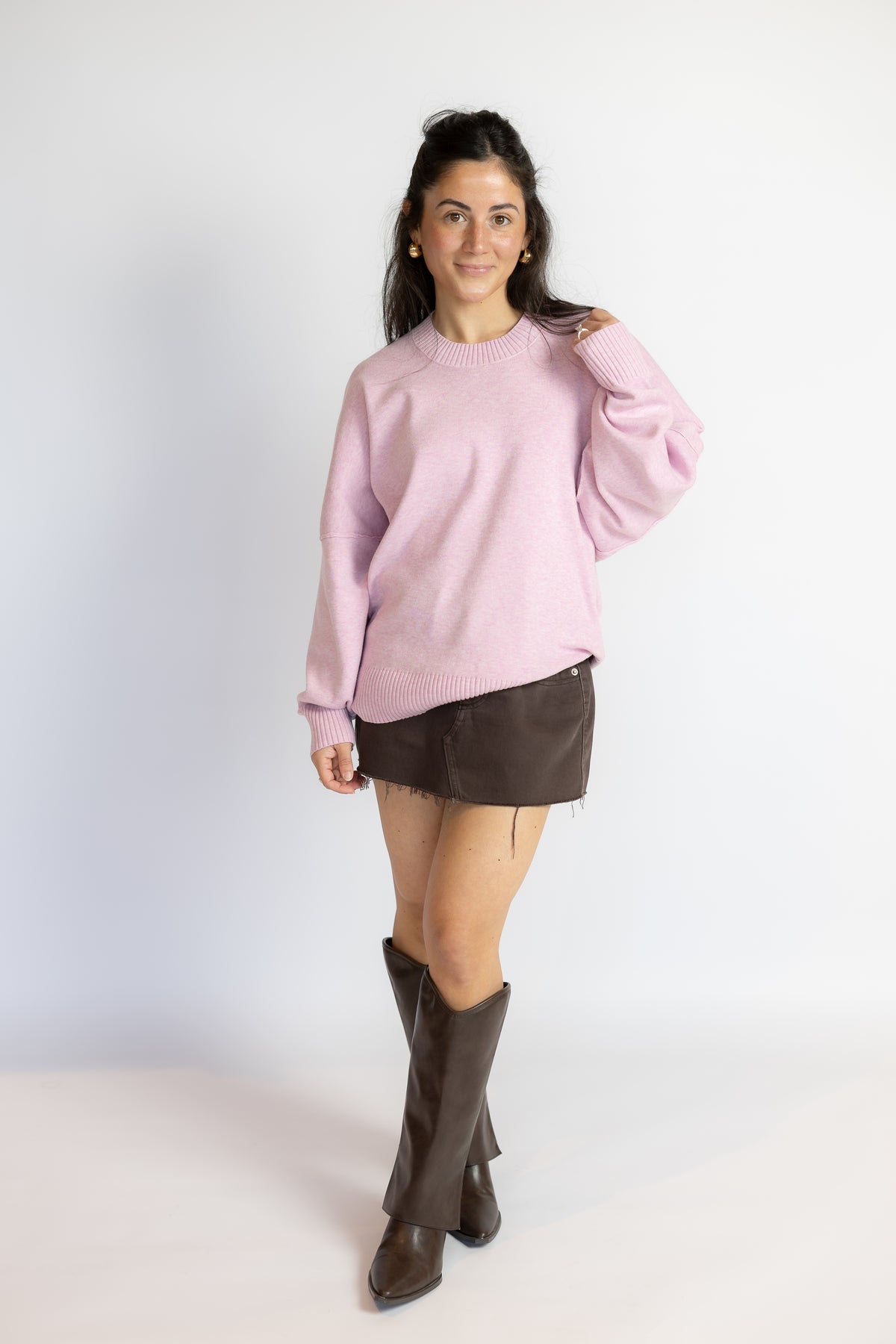 A woman modeling a light pink crewneck sweatshirt, a dark brown mini skirt, and dark brown knee-high boots, standing against a plain white studio background.