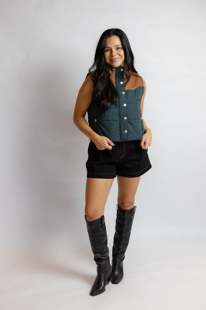 A woman modeling a sleeveless button-front vest in dark teal and black with contrast stitching, paired with black shorts and tall black boots, against a light gray studio background.