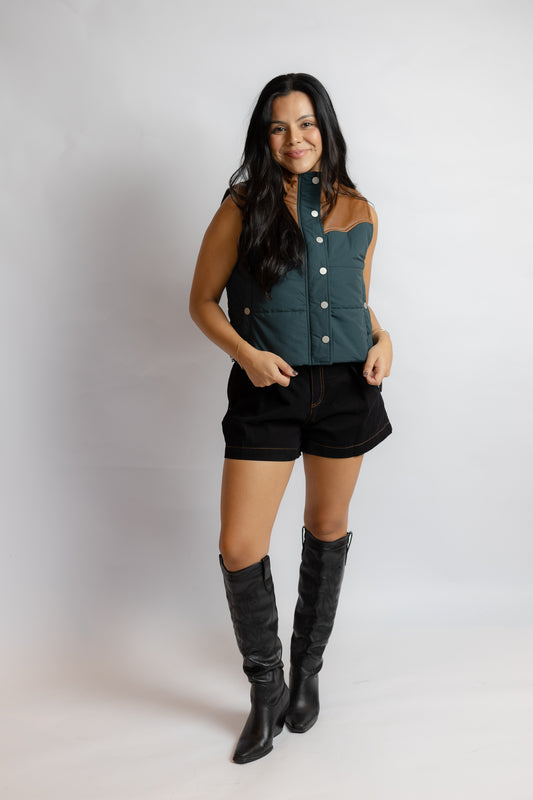 A woman modeling a sleeveless button-front vest in dark teal and black with contrast stitching, paired with black shorts and tall black boots, against a light gray studio background.
