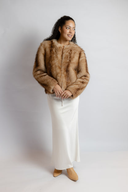 A woman modeling a cropped brown fur jacket over a long white satin skirt, standing against a light gray studio background.