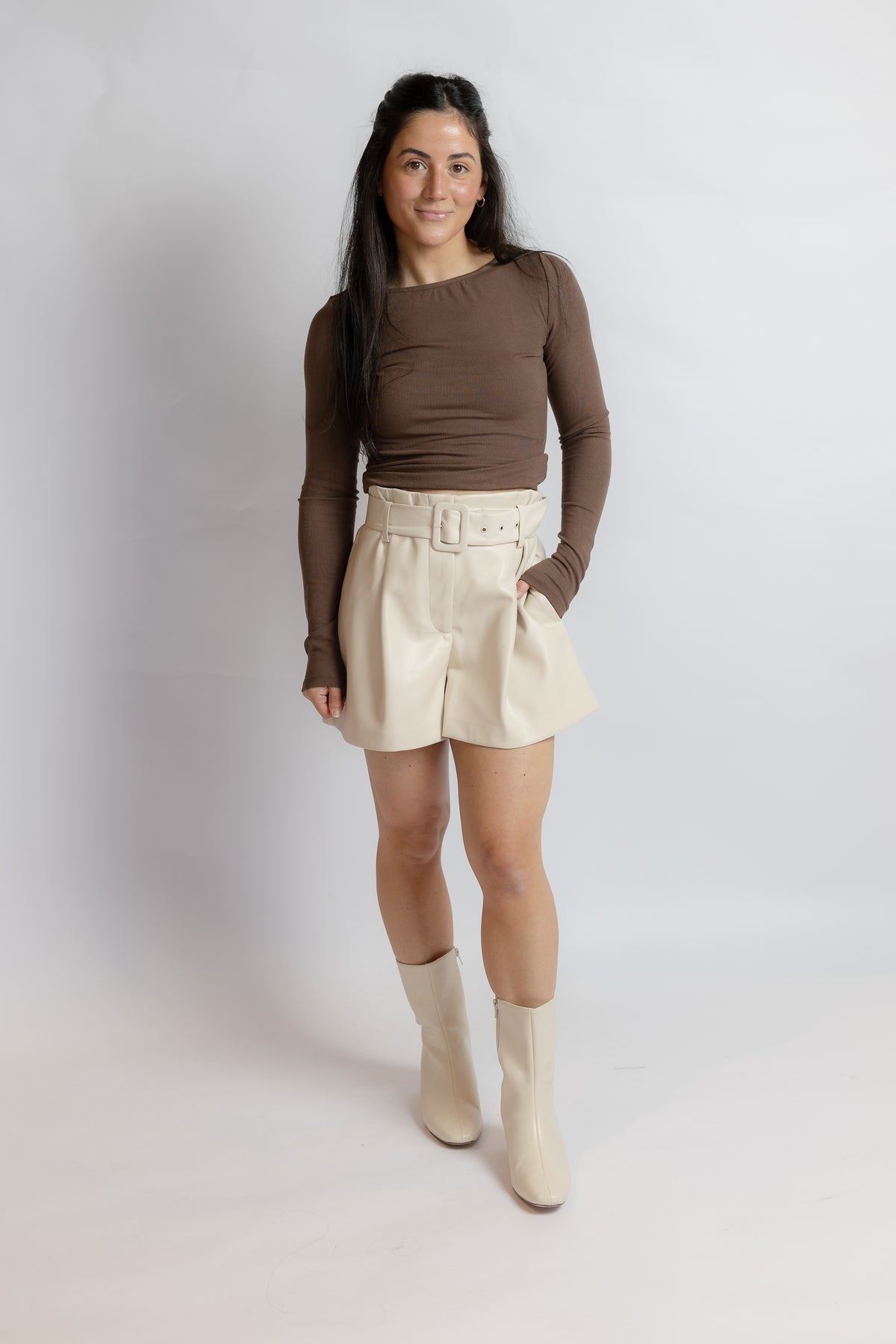 A woman modeling a brown long-sleeve ribbed top and cream high-waisted shorts with matching belt, paired with cream ankle boots, against a light gray studio background.