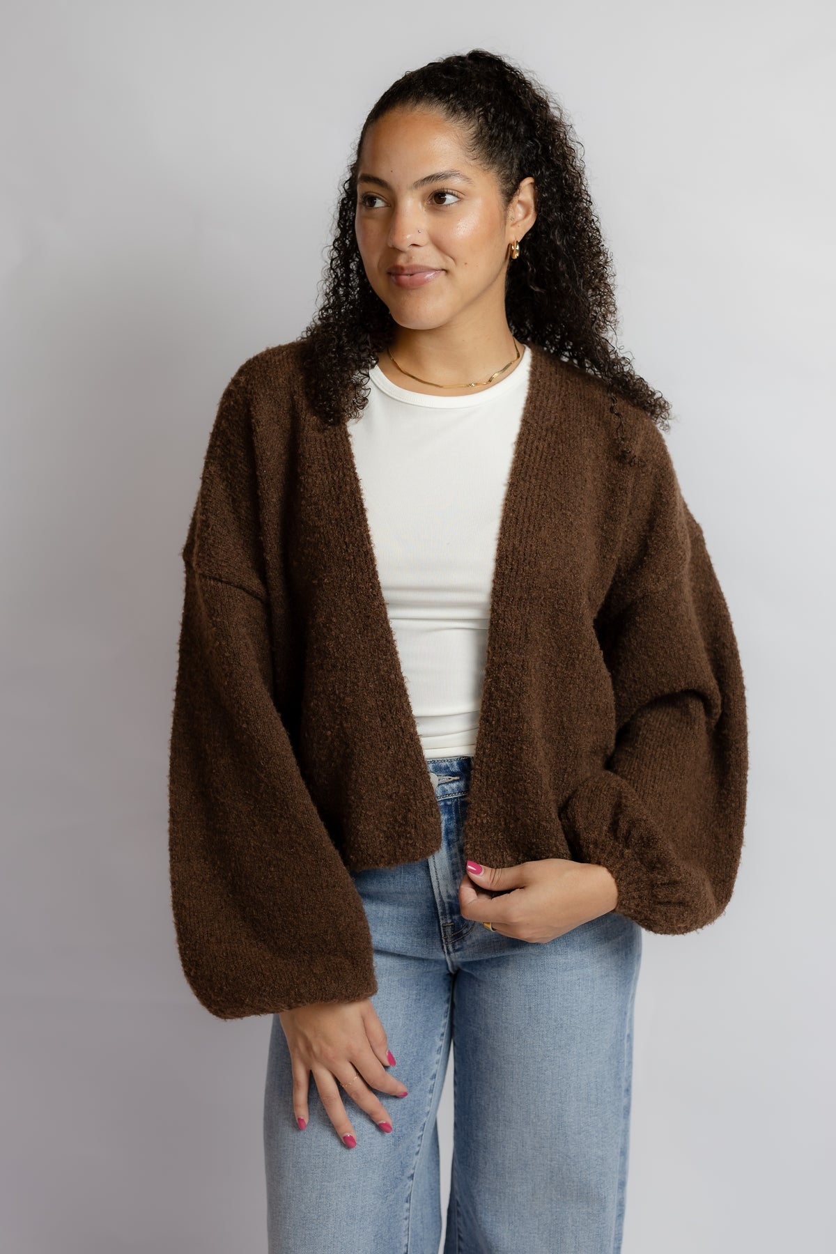 A woman modeling a brown fuzzy open-front cardigan over a white top and light blue jeans, photographed against a plain light gray studio background.