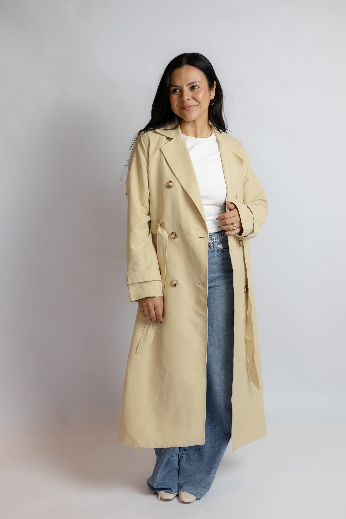 A woman modeling a long beige trench coat over a white top and blue jeans, standing against a plain light gray studio background.