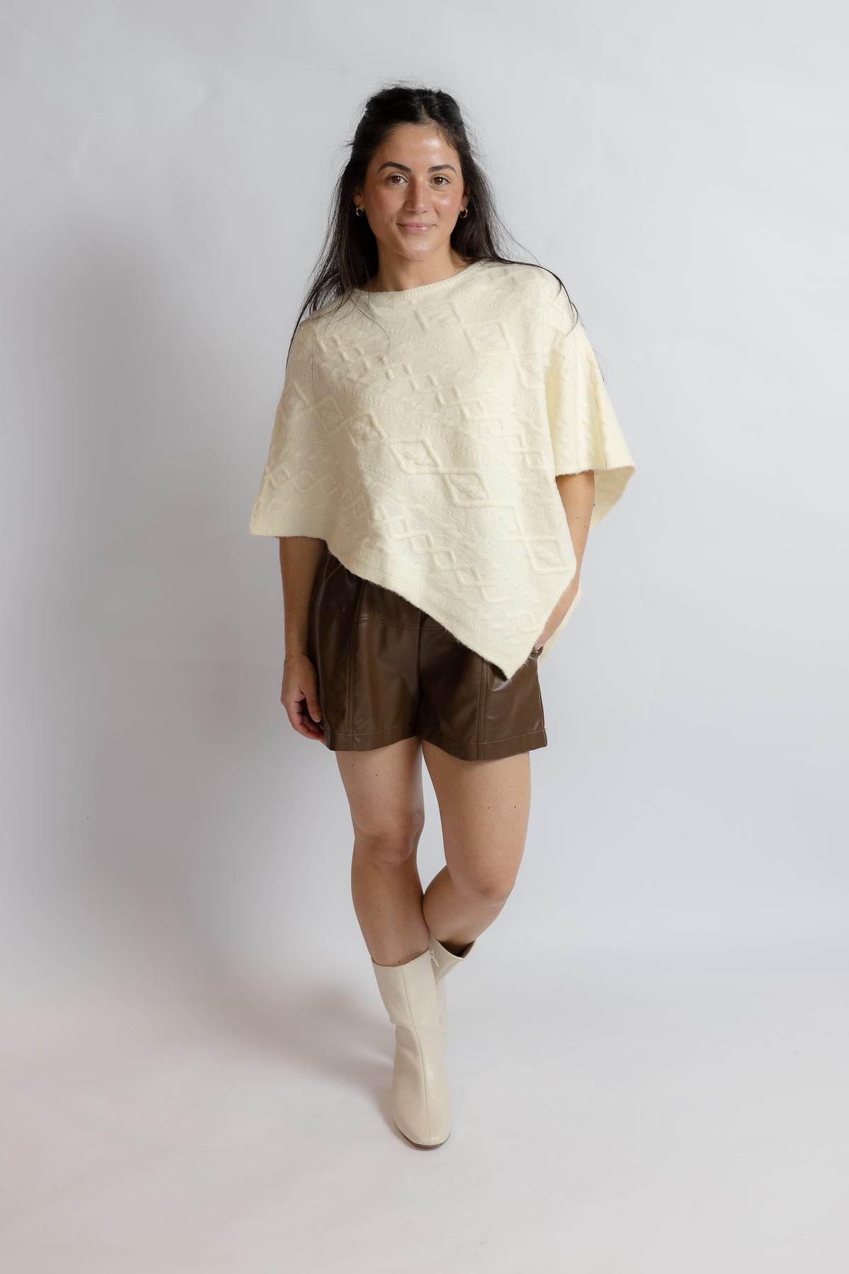 A woman modeling a cream-colored textured short-sleeve poncho top paired with brown faux-leather shorts and cream ankle boots, standing against a light gray studio background.