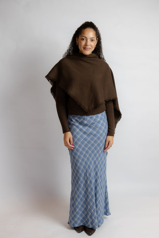 A woman modeling a dark brown fringed wrap shawl over a brown long-sleeve top and a blue and white argyle patterned maxi skirt, standing against a light gray studio background.