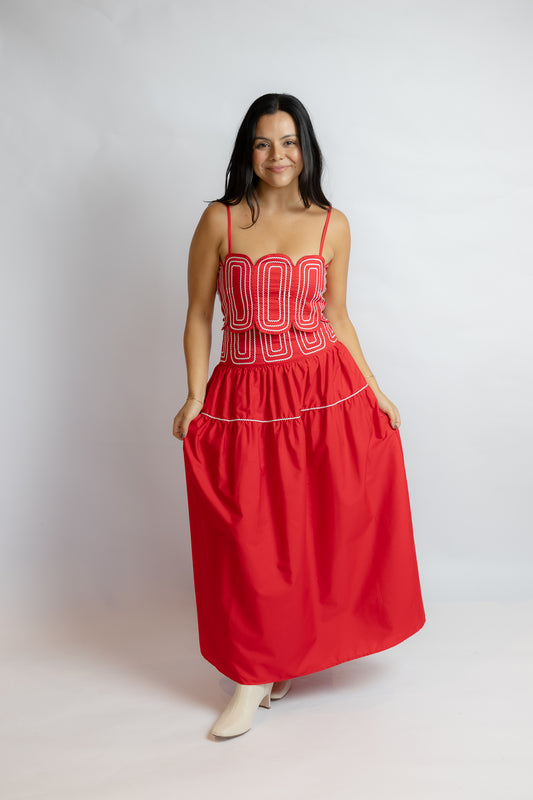 A woman modeling a bright red maxi dress with a structured bodice featuring white embroidered lettering, thin spaghetti straps, and a gathered skirt with side pockets, set against a plain light gray studio background.