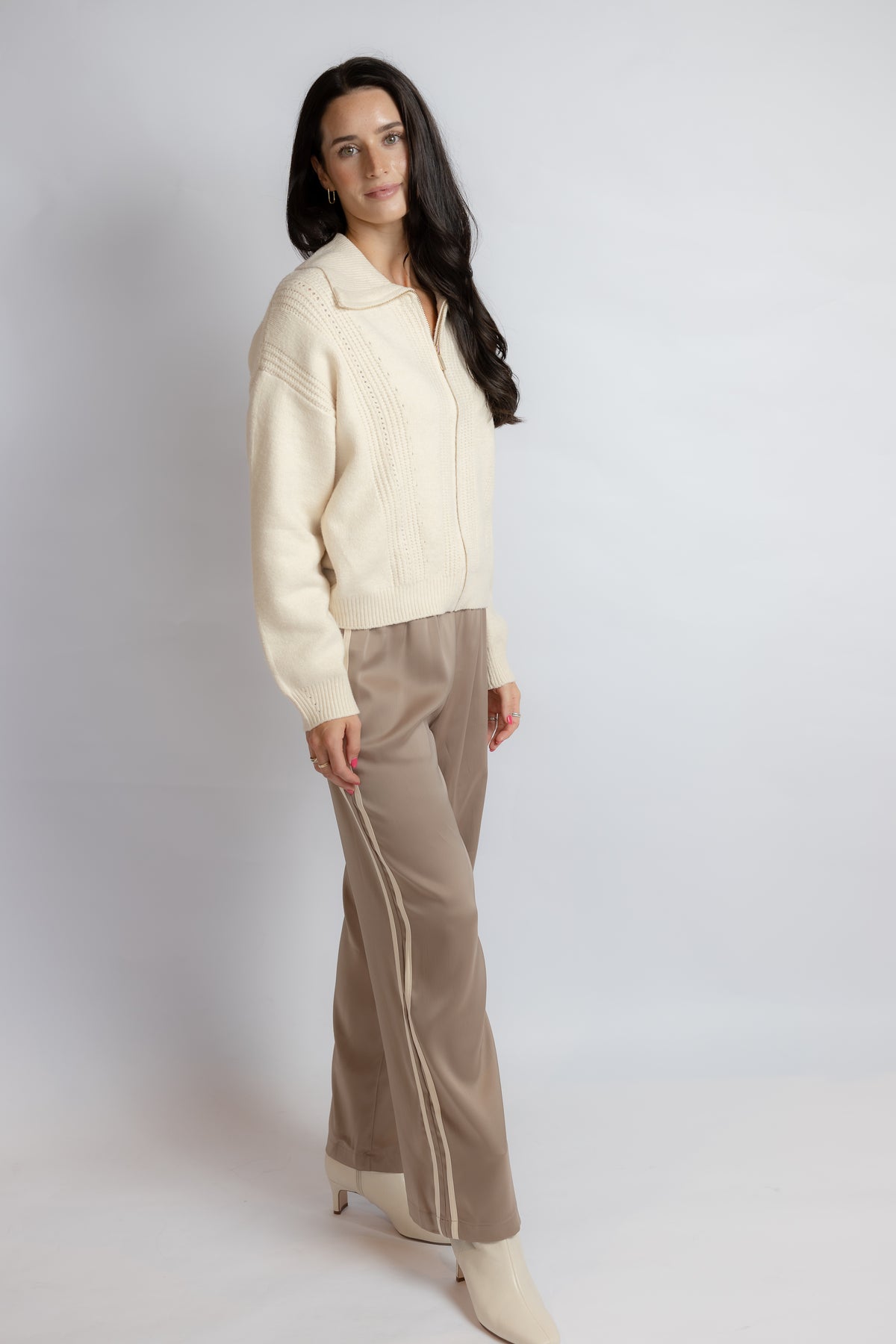 A woman modeling a cream ribbed knit zip-up cardigan and taupe wide-leg trousers with white side stripes, standing against a light gray studio background.