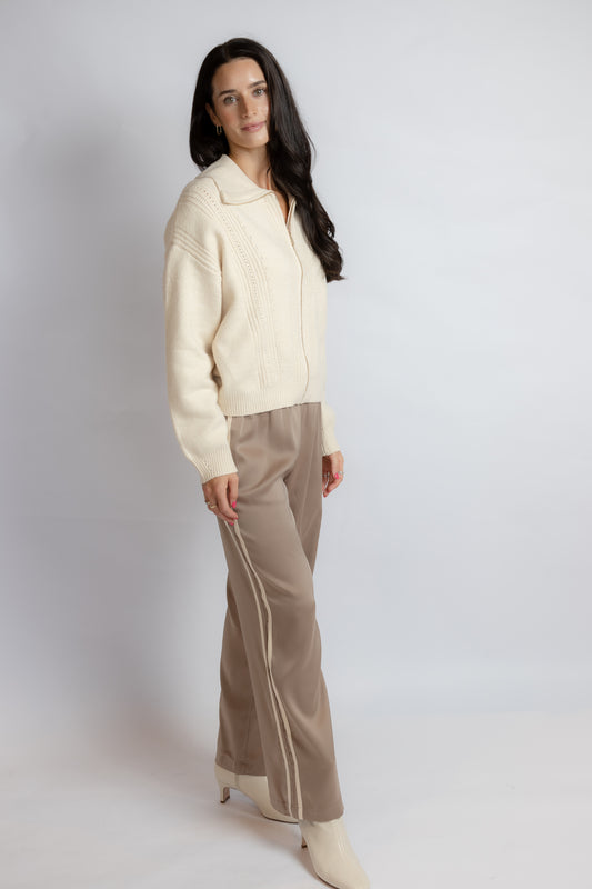 A woman modeling a cream ribbed knit zip-up cardigan and taupe wide-leg trousers with white side stripes, standing against a light gray studio background.
