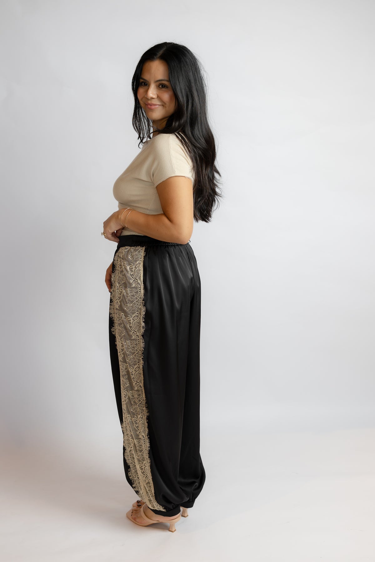 A woman modeling a pair of black wide-leg pants with a beige patterned side panel, paired with a beige short-sleeve top, against a plain light gray studio background.
