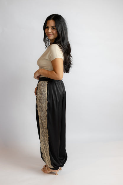 A woman modeling a pair of black wide-leg pants with a beige patterned side panel, paired with a beige short-sleeve top, against a plain light gray studio background.