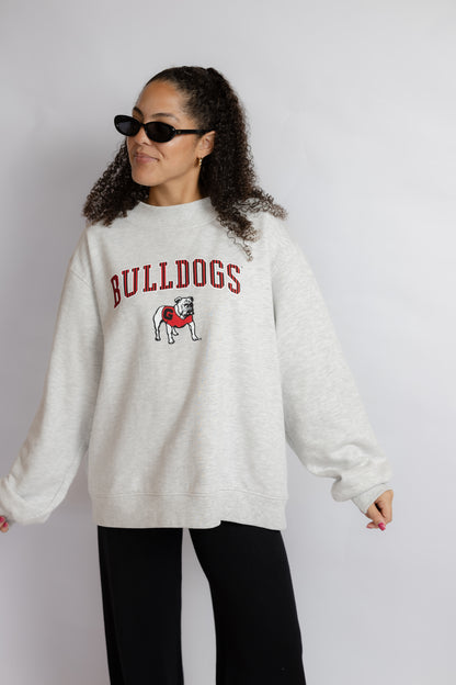 A woman modeling an oversized off-white heather sweatshirt with 'BULLDOGS' and a bulldog graphic in red and black on the chest, paired with black pants against a light gray studio background.