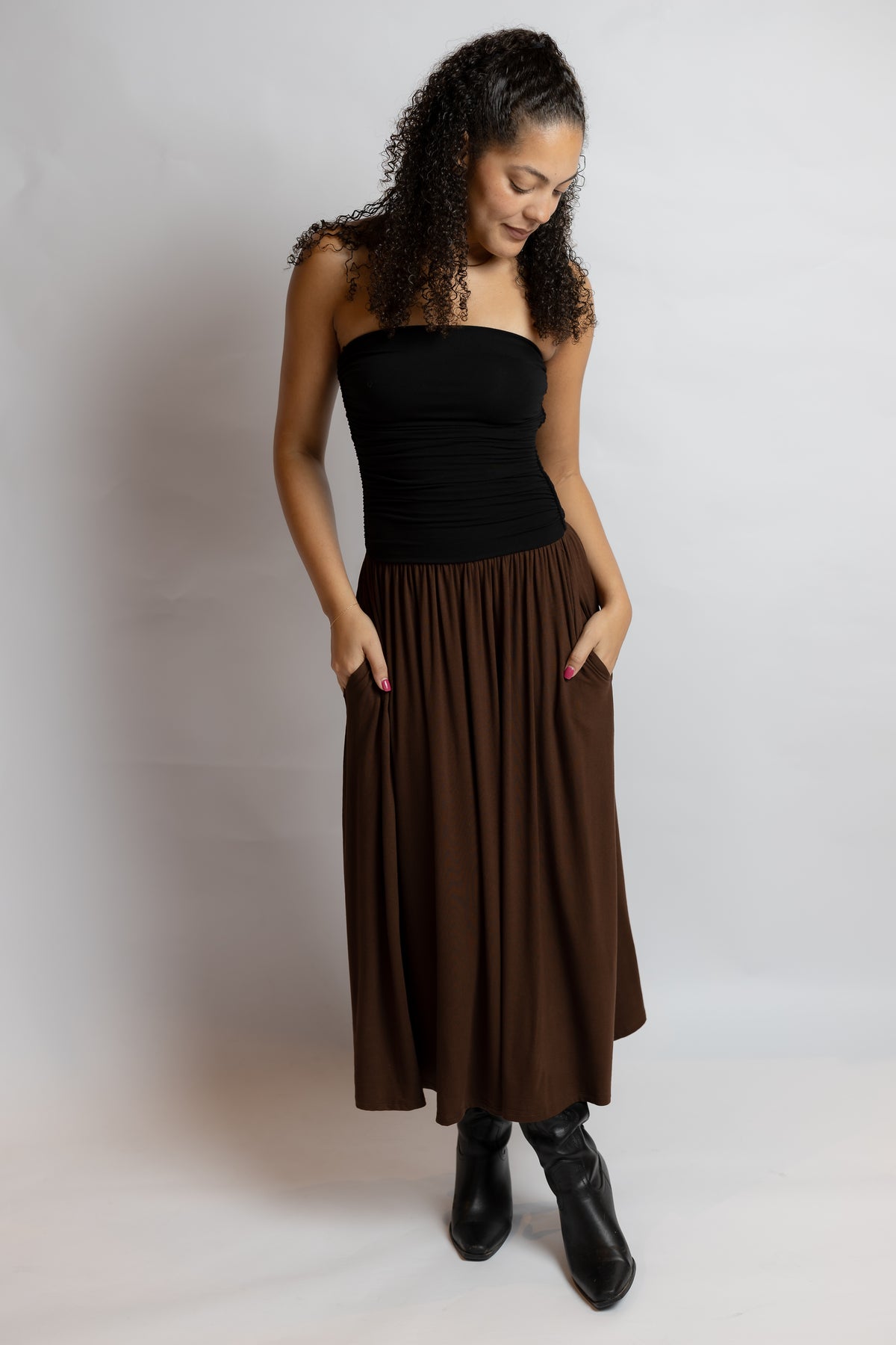 A woman modeling a dark brown pleated midi skirt with side pockets, paired with a black strapless top and black ankle boots, standing against a light gray studio background.