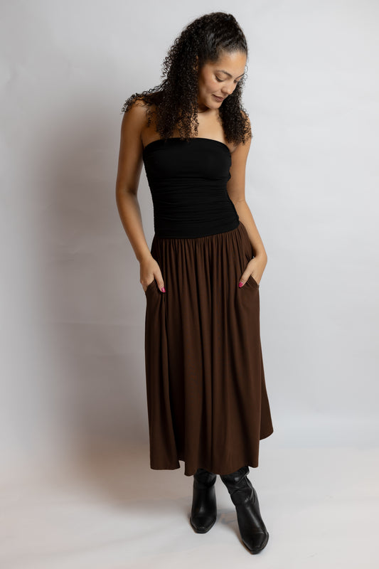 A woman modeling a dark brown pleated midi skirt with side pockets, paired with a black strapless top and black ankle boots, standing against a light gray studio background.