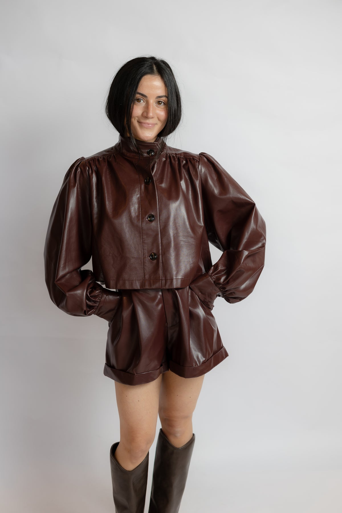A woman modeling a shiny dark brown faux leather two-piece set consisting of a cropped long-sleeve jacket with puffed sleeves and matching shorts, paired with dark brown boots, against a light gray background.
