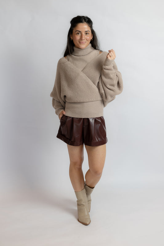 A woman modeling a beige knit turtleneck sweater with draped sleeves, paired with dark brown faux-leather shorts and beige ankle boots, against a light gray studio background.