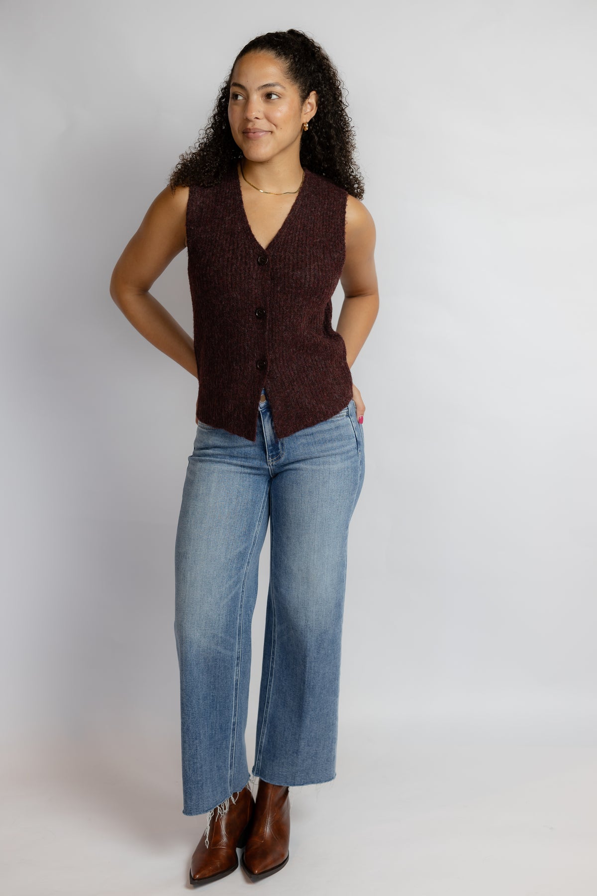 A woman modeling a sleeveless, v-neck knit vest in a deep burgundy brown, paired with light-wash blue jeans and brown leather boots, against a plain light gray studio background.