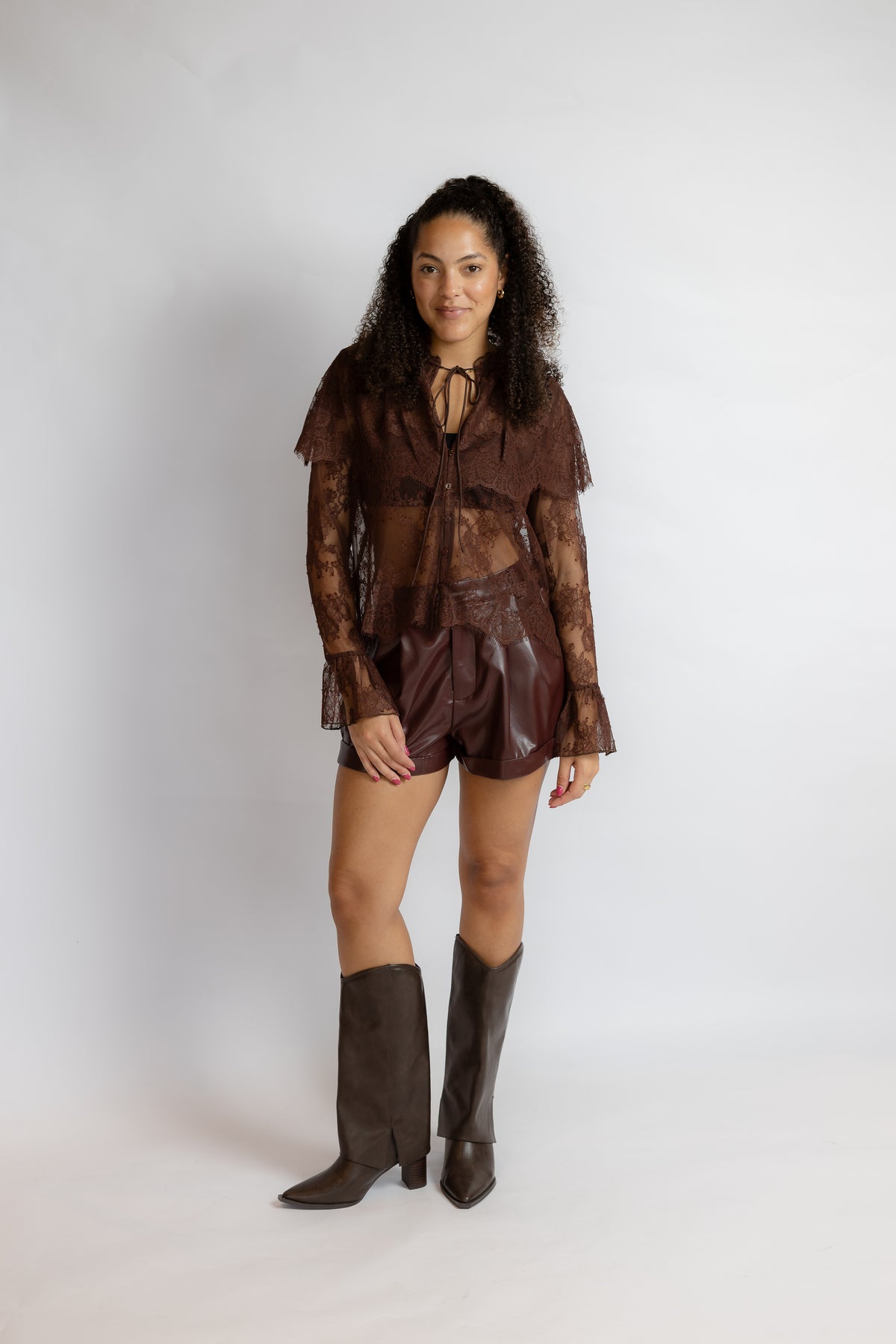 A woman modeling a brown lace and faux-leather outfit consisting of a long-sleeve ruffled top, matching shorts, and dark brown knee-high boots, photographed against a plain light gray studio background.