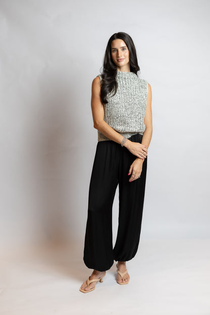 A woman modeling a sleeveless knit top with a textured silver and black pattern, paired with black wide-leg pants and beige sandals, standing against a light gray studio backdrop.