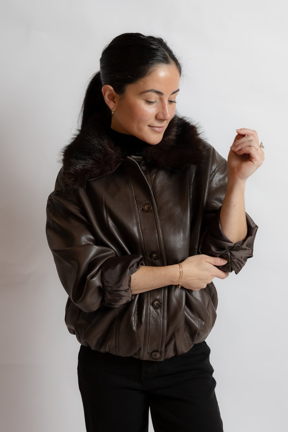 A woman wearing a dark brown faux-leather jacket with a dark brown fur collar, paired with black pants, photographed against a light gray studio background.