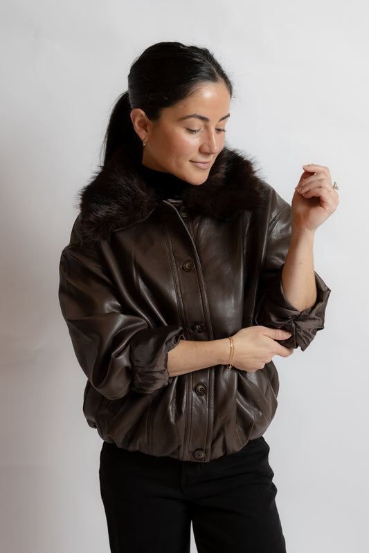 A woman wearing a dark brown faux-leather jacket with a dark brown fur collar, paired with black pants, photographed against a light gray studio background.