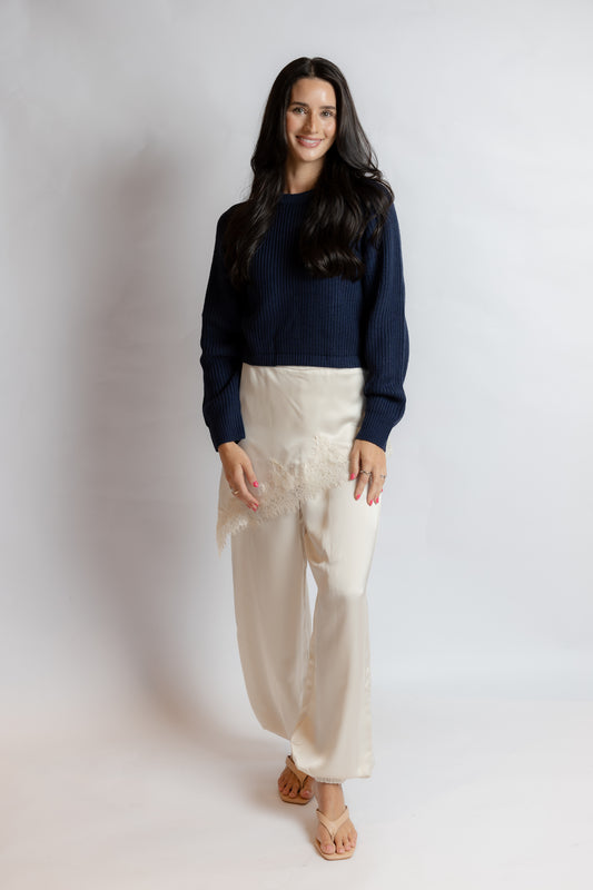A woman modeling a dark navy blue knit sweater paired with cream wide-leg trousers featuring a lace detail, standing against a light gray studio background.