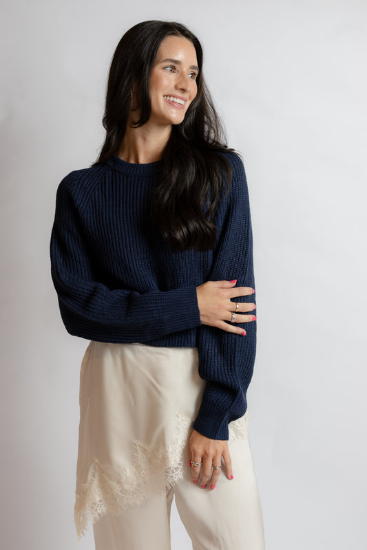 A woman wearing a navy blue ribbed knit sweater and cream pants with lace trim, standing against a light gray studio background.