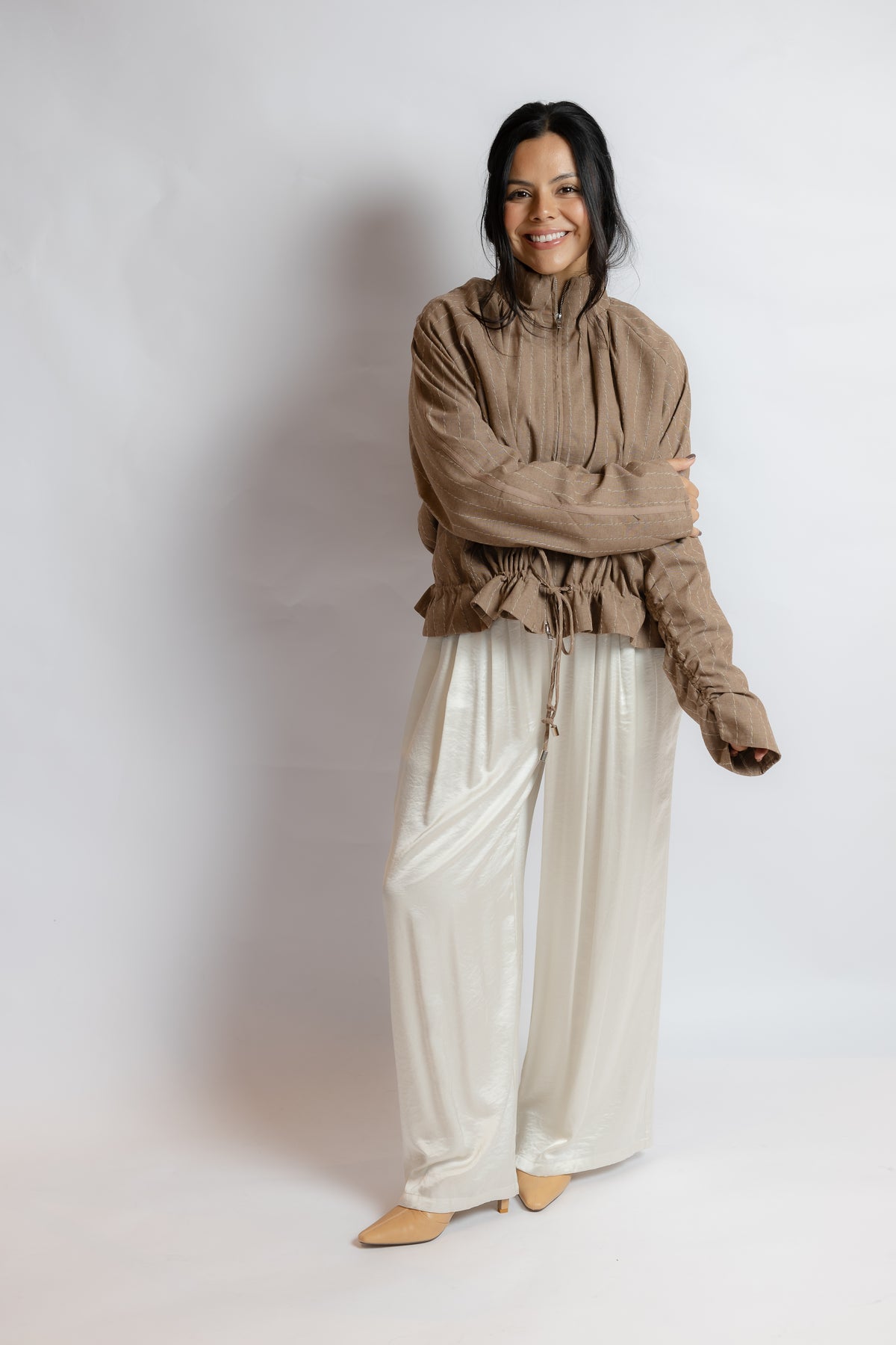 A woman modeling a brown ruffled long-sleeve blouse and shiny cream wide-leg pants against a light gray studio background.