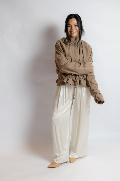 A woman modeling a brown ruffled long-sleeve blouse and shiny cream wide-leg pants against a light gray studio background.