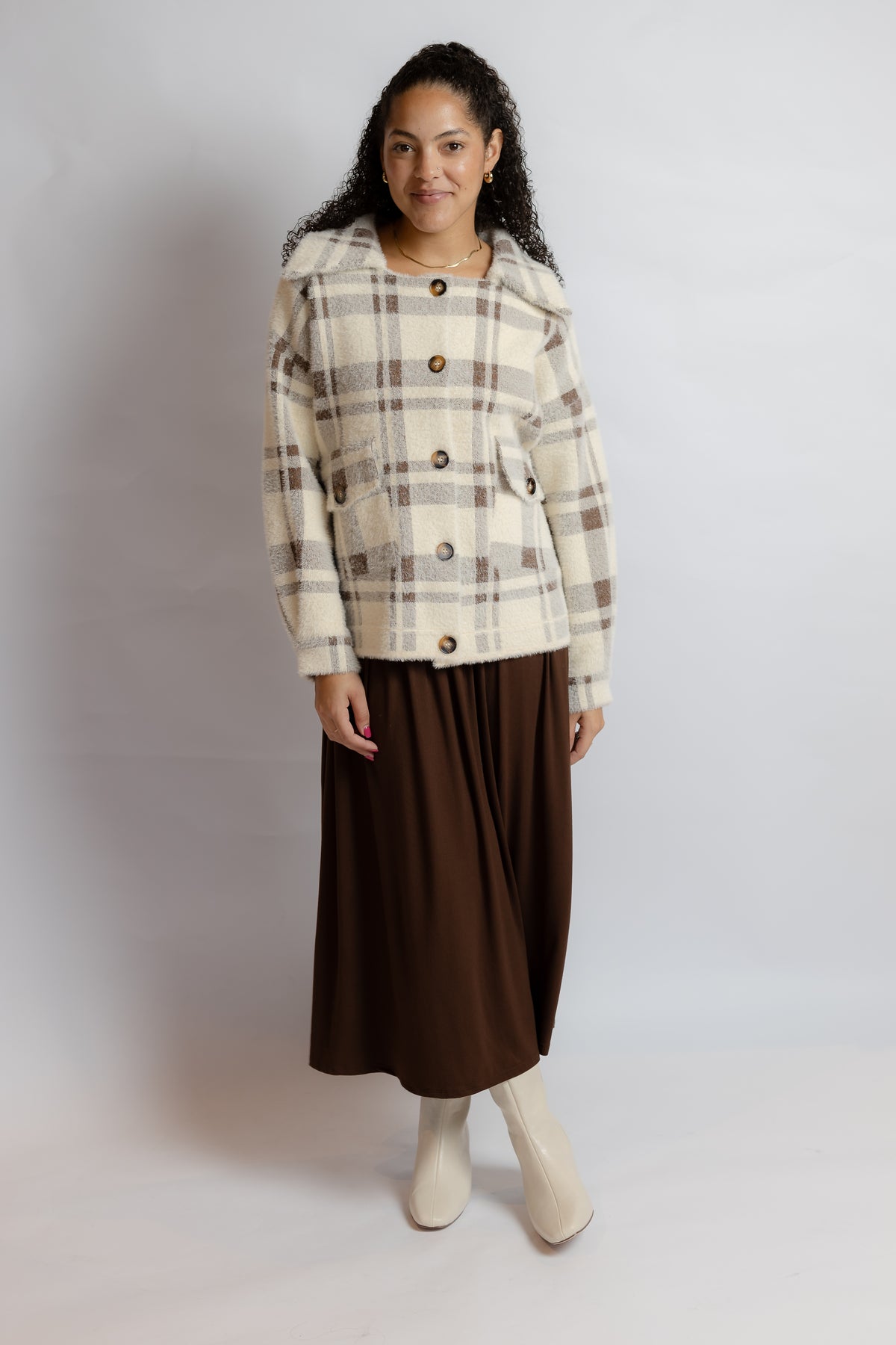A woman modeling a cream and brown plaid cropped jacket with large buttons, paired with a dark brown midi skirt and off-white boots, against a light gray studio background.