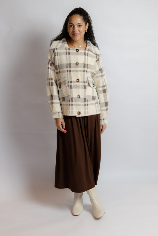 A woman modeling a cream and brown plaid cropped jacket with large buttons, paired with a dark brown midi skirt and off-white boots, against a light gray studio background.