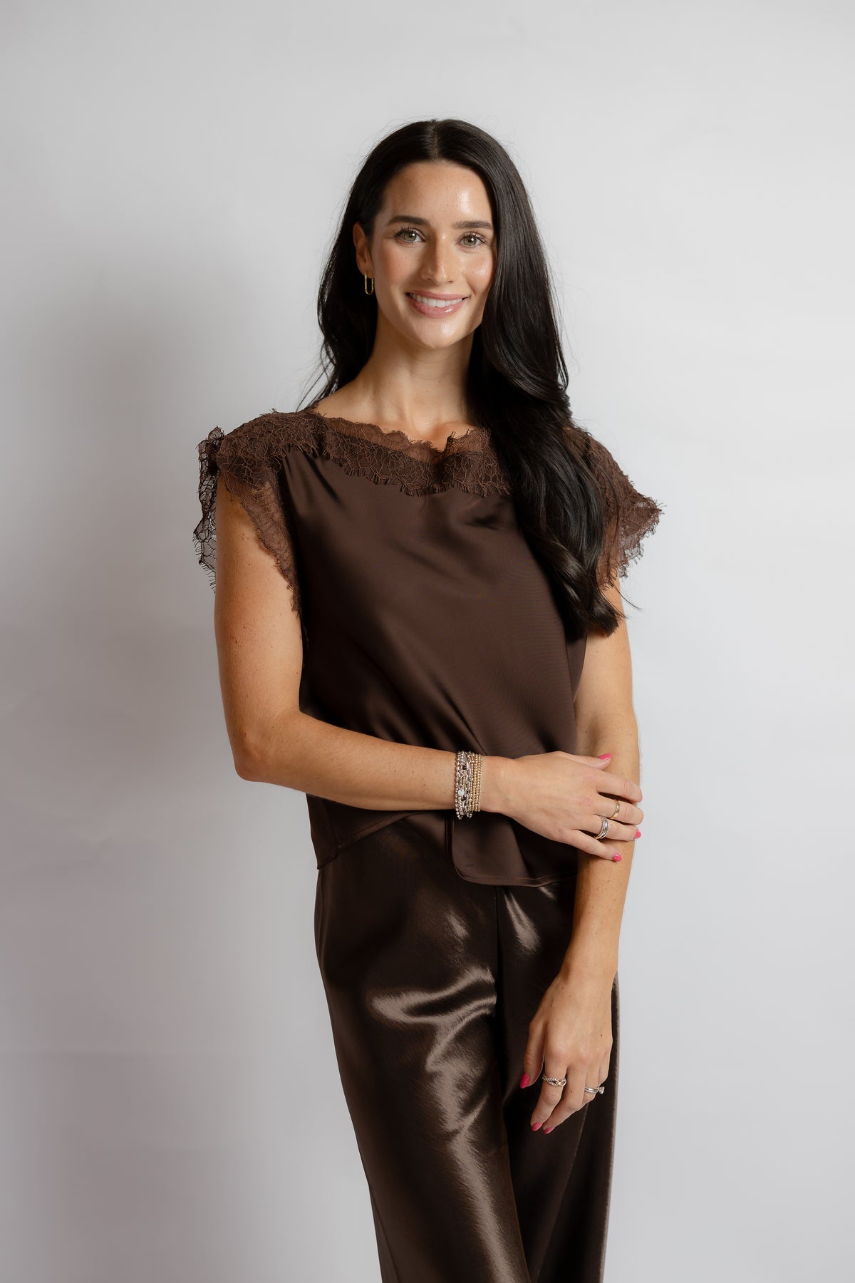 A woman modeling a brown satin-like jumpsuit with sheer lace cap sleeves and a matching headband, standing against a light gray studio background.
